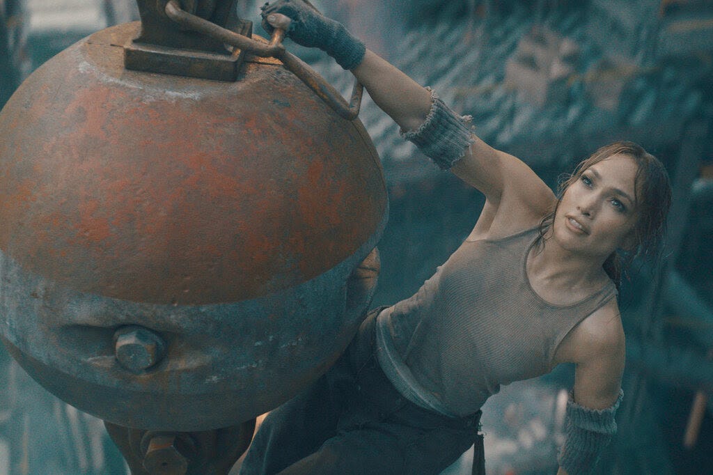 A still of Jennifer Lopez in a taupe tank top hanging by one arm from a wrecking ball.